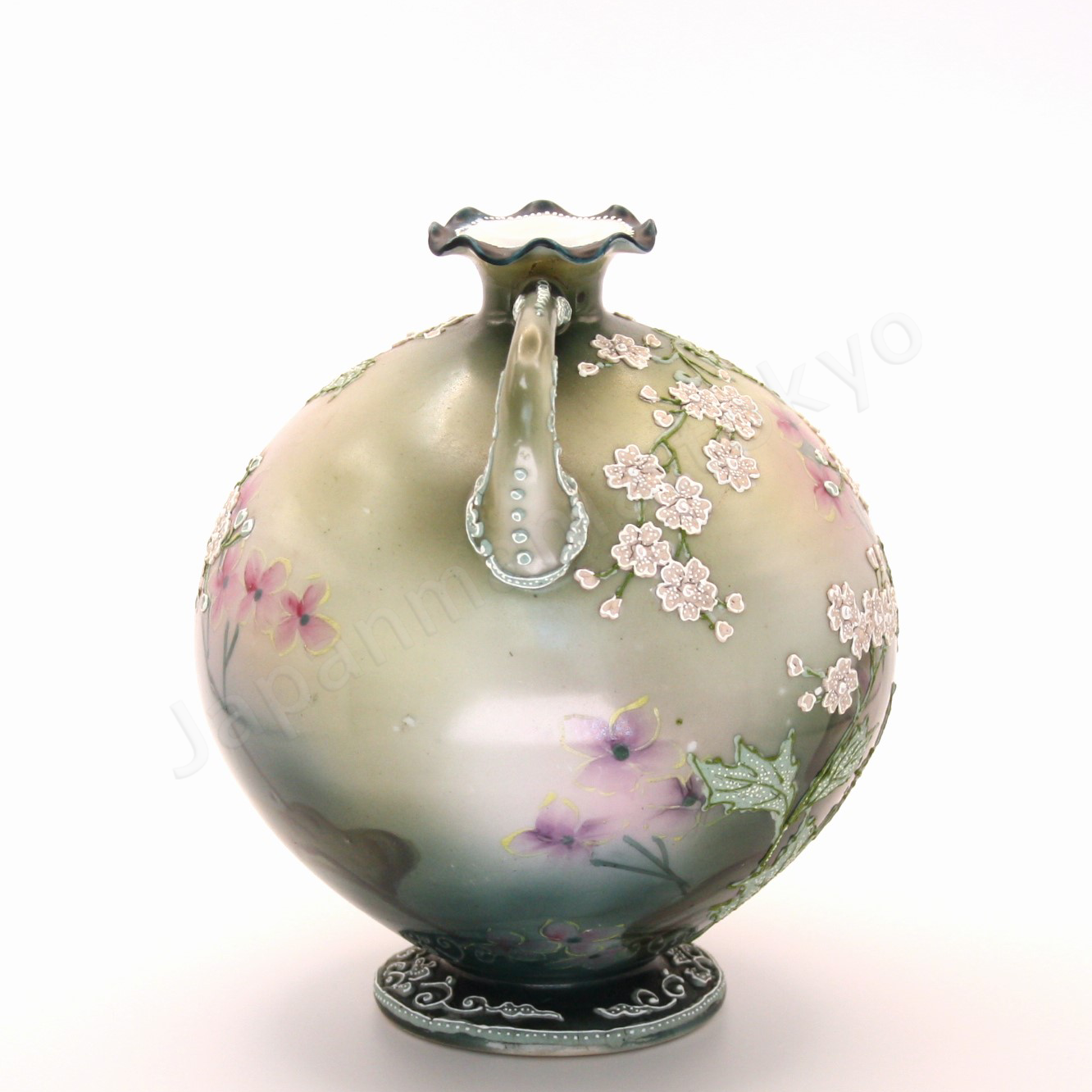 Side view: Japanese old fine china. Old Noritake.
This exquisite and captivating piece showcases polychrome and Moriage decoration, featuring delicate white flowers tinged with soft pink and leaves outlined in green. The upper and lower portions are finished in a smoky green, while the center glows with a radiant brightness, as though illuminated by light. The elegantly small mouth of the base, complemented by graceful handles on both sides, adds to its charm and timeless appeal.