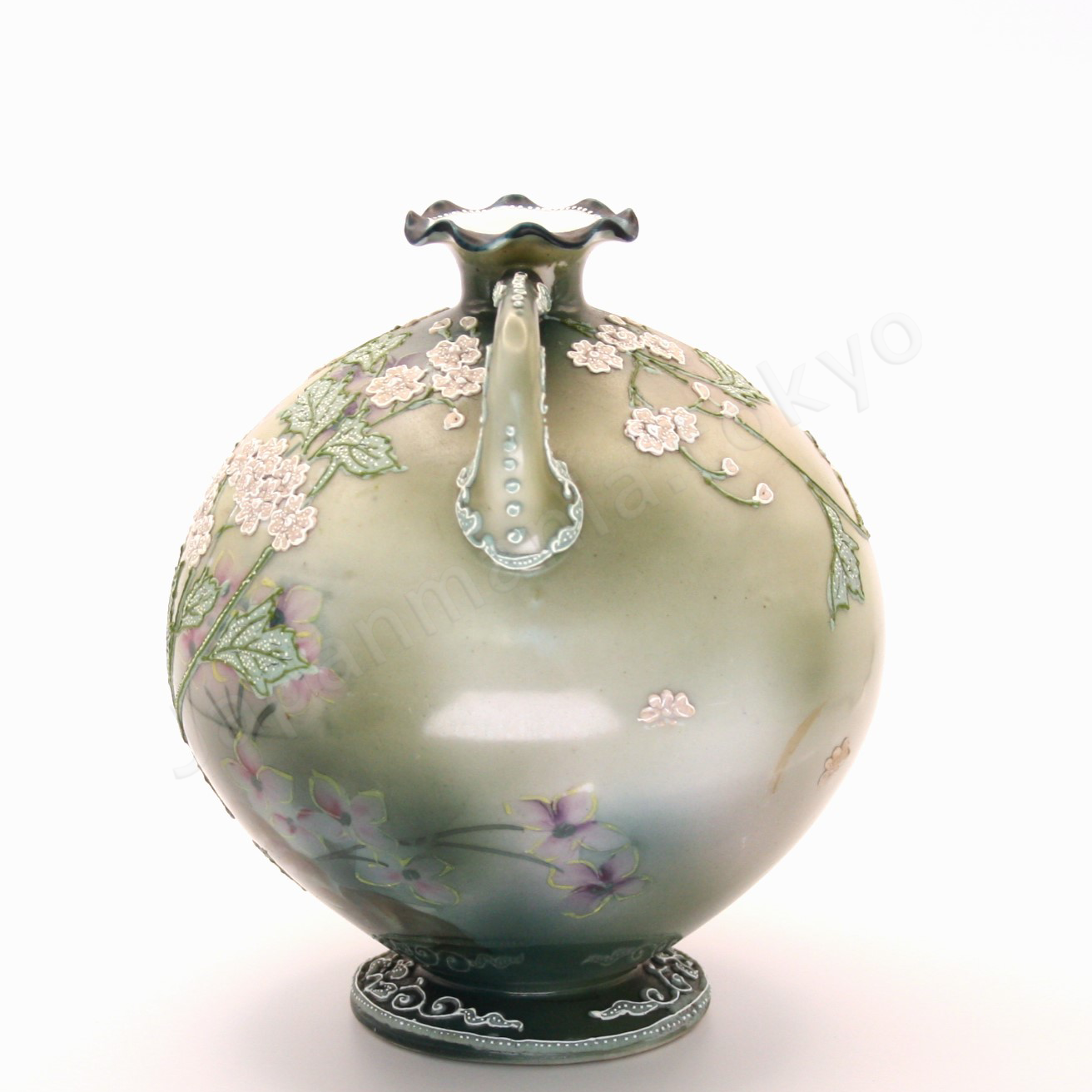 Side view: Japanese old fine china. Old Noritake.
This exquisite and captivating piece showcases polychrome and Moriage decoration, featuring delicate white flowers tinged with soft pink and leaves outlined in green. The upper and lower portions are finished in a smoky green, while the center glows with a radiant brightness, as though illuminated by light. The elegantly small mouth of the base, complemented by graceful handles on both sides, adds to its charm and timeless appeal.