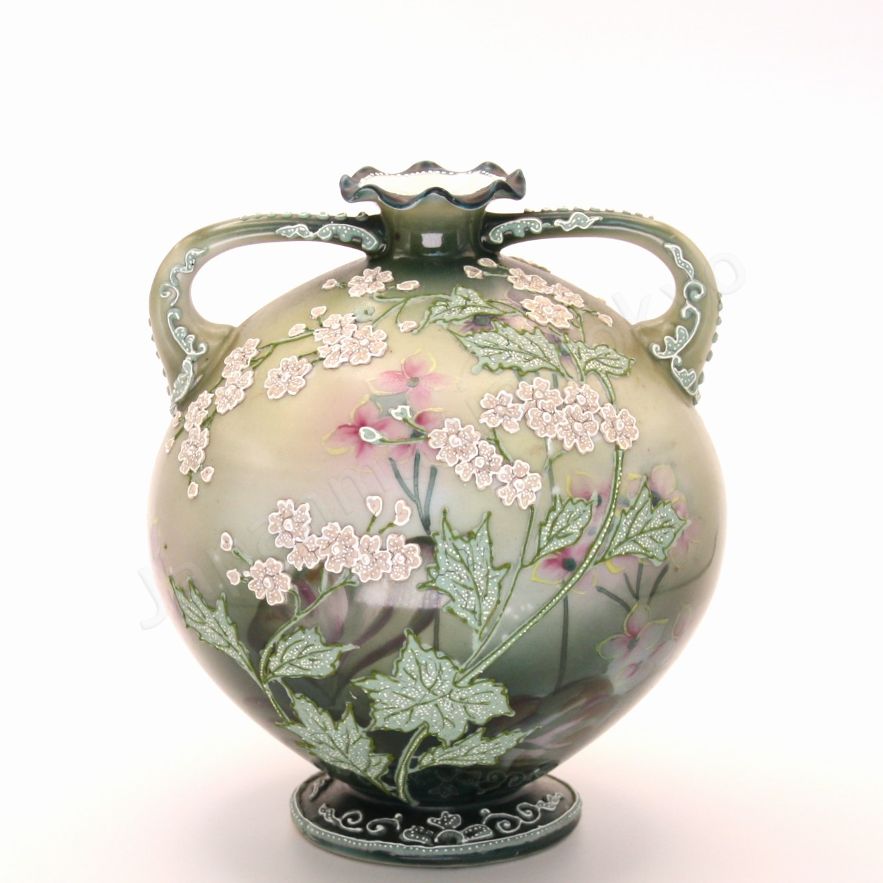 Front view: Japanese old fine china. Old Noritake.
This exquisite and captivating piece showcases polychrome and Moriage decoration, featuring delicate white flowers tinged with soft pink and leaves outlined in green. The upper and lower portions are finished in a smoky green, while the center glows with a radiant brightness, as though illuminated by light. The elegantly small mouth of the base, complemented by graceful handles on both sides, adds to its charm and timeless appeal.