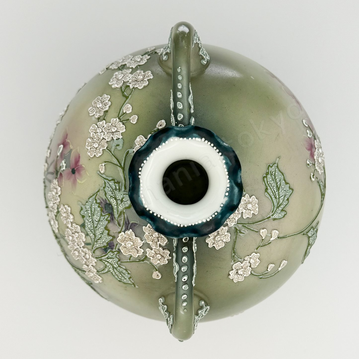 Top view: Japanese old fine china. Old Noritake.
This exquisite and captivating piece showcases polychrome and Moriage decoration, featuring delicate white flowers tinged with soft pink and leaves outlined in green. The upper and lower portions are finished in a smoky green, while the center glows with a radiant brightness, as though illuminated by light. The elegantly small mouth of the base, complemented by graceful handles on both sides, adds to its charm and timeless appeal.