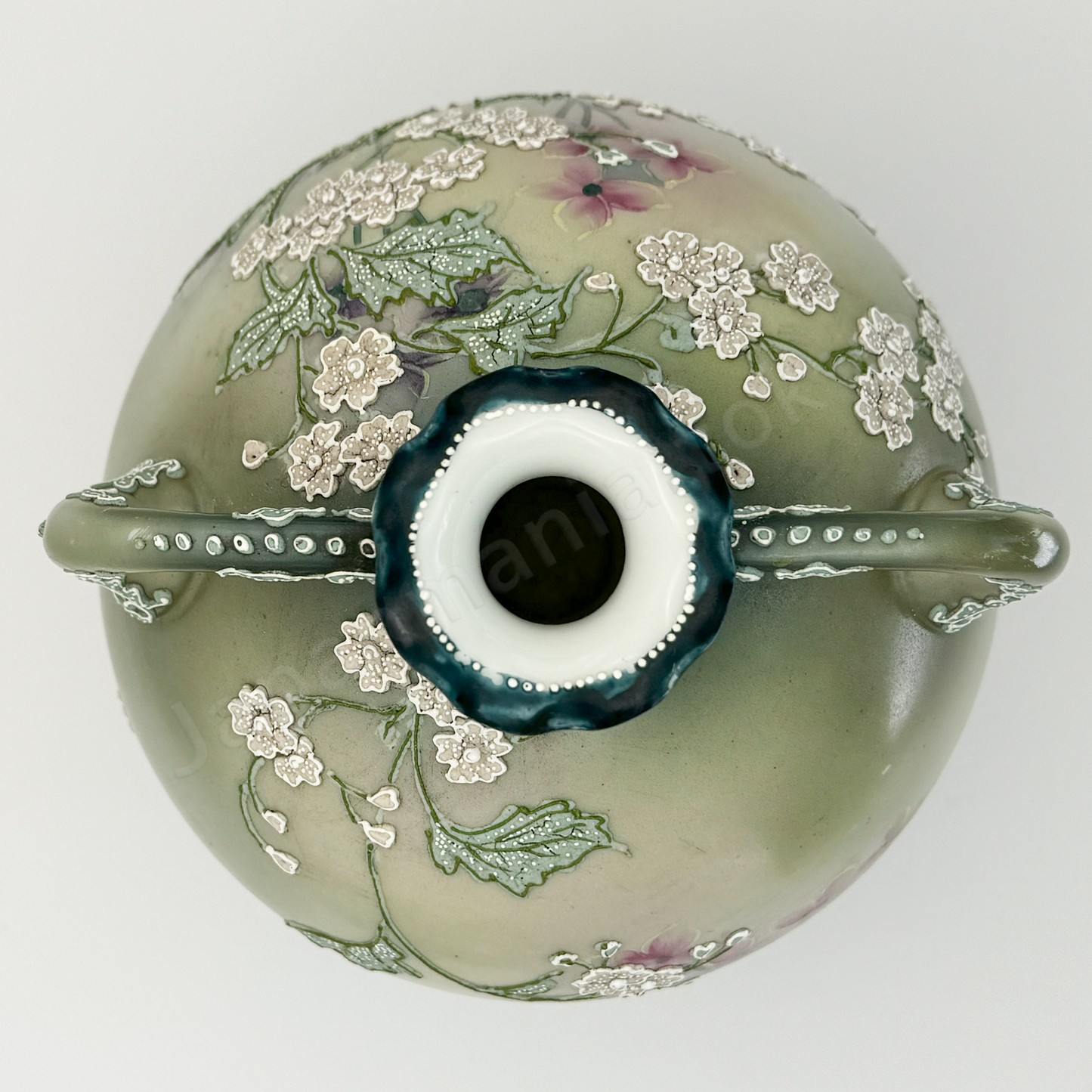 Top view: Japanese old fine china. Old Noritake.
This exquisite and captivating piece showcases polychrome and Moriage decoration, featuring delicate white flowers tinged with soft pink and leaves outlined in green. The upper and lower portions are finished in a smoky green, while the center glows with a radiant brightness, as though illuminated by light. The elegantly small mouth of the base, complemented by graceful handles on both sides, adds to its charm and timeless appeal.